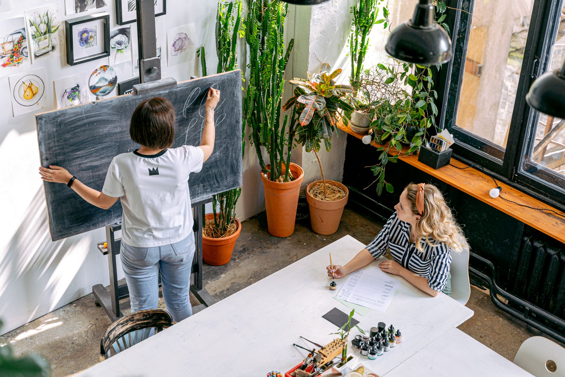 Art workshop with instructor at blackboard and student in studio surrounded by plants.