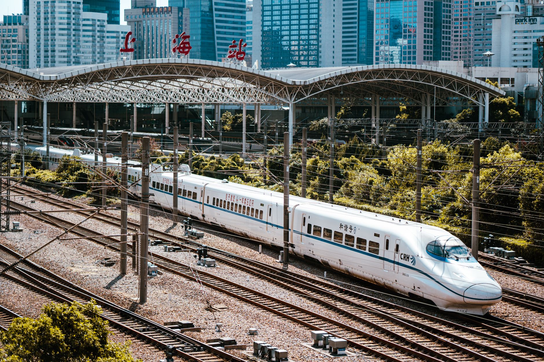 A high-speed train passing through Shanghai Railway Station with city skyline in the background.
