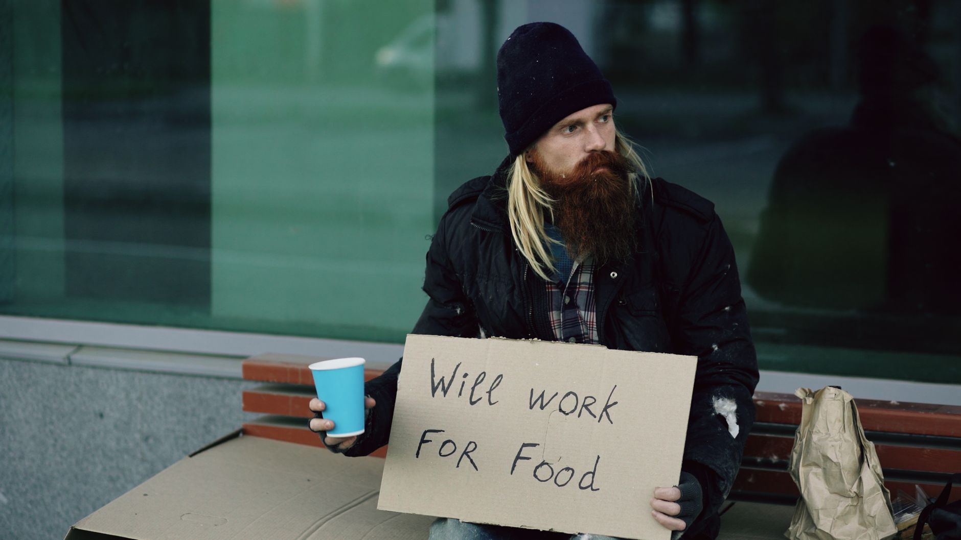 Homeless man with sign "Will Work for Food" on a city street bench.