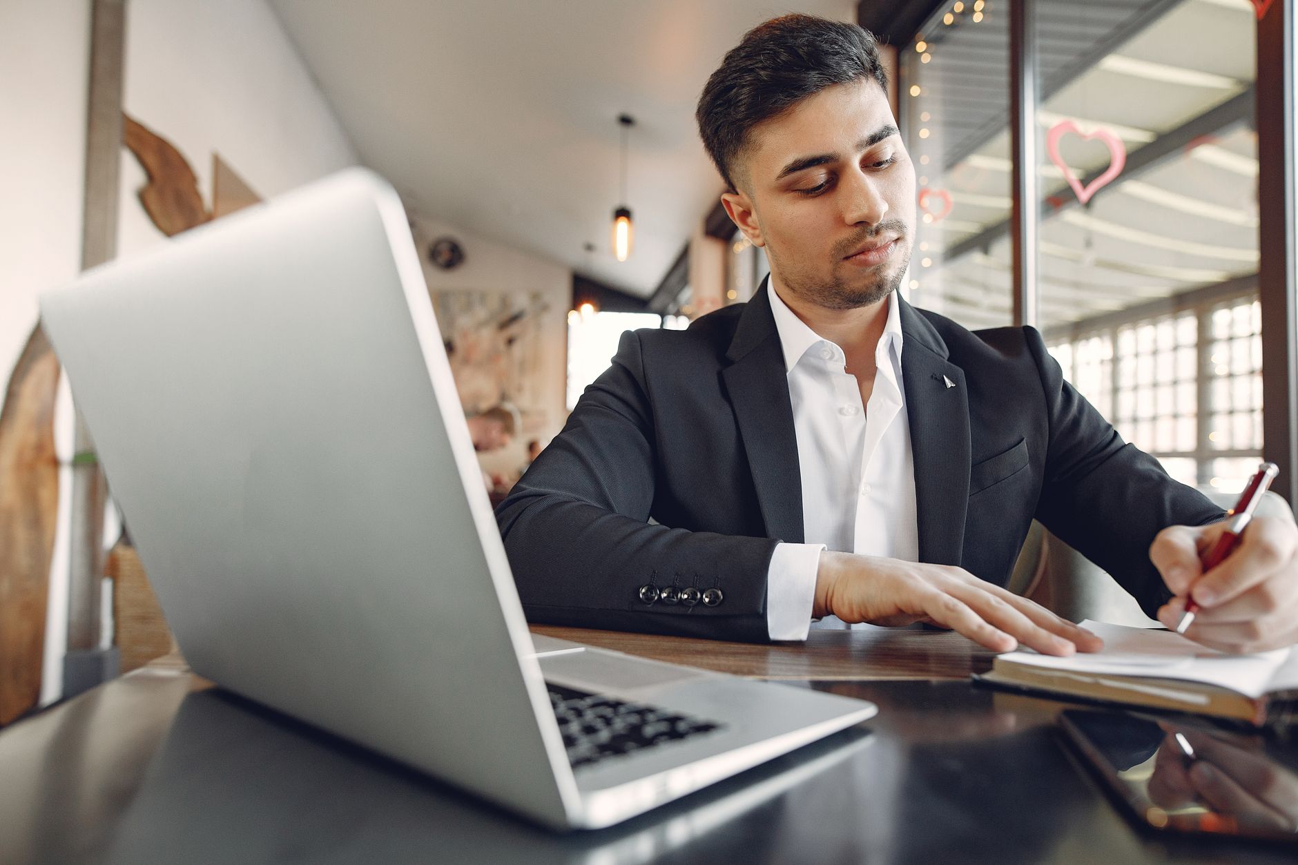 From below of concentrated ethnic male in formal wear sitting in creative workplace and taking notes while using laptop and working on busin