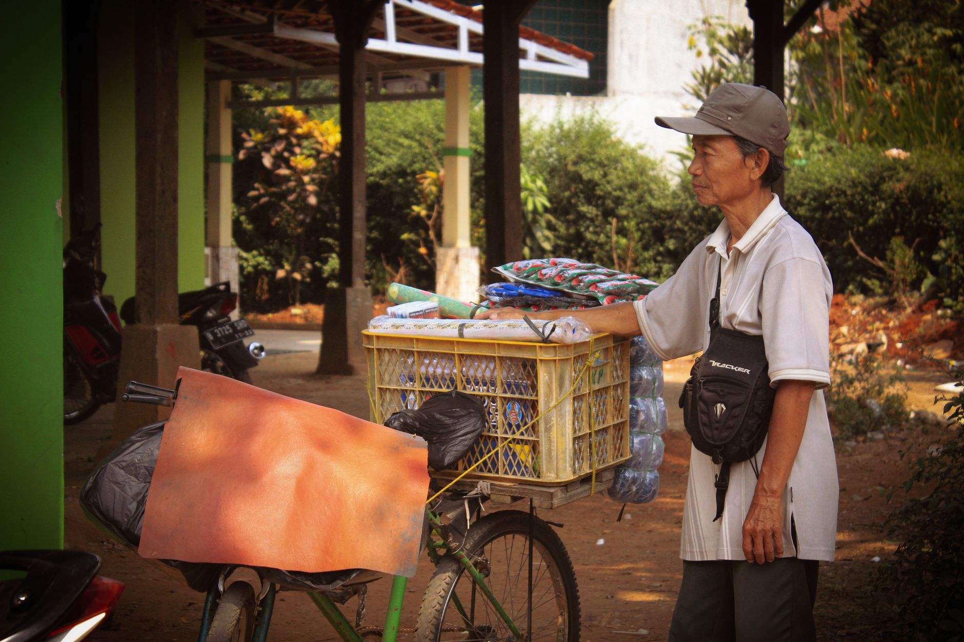 A man standing beside a bicycle loaded with goods, outdoors in a shaded area.