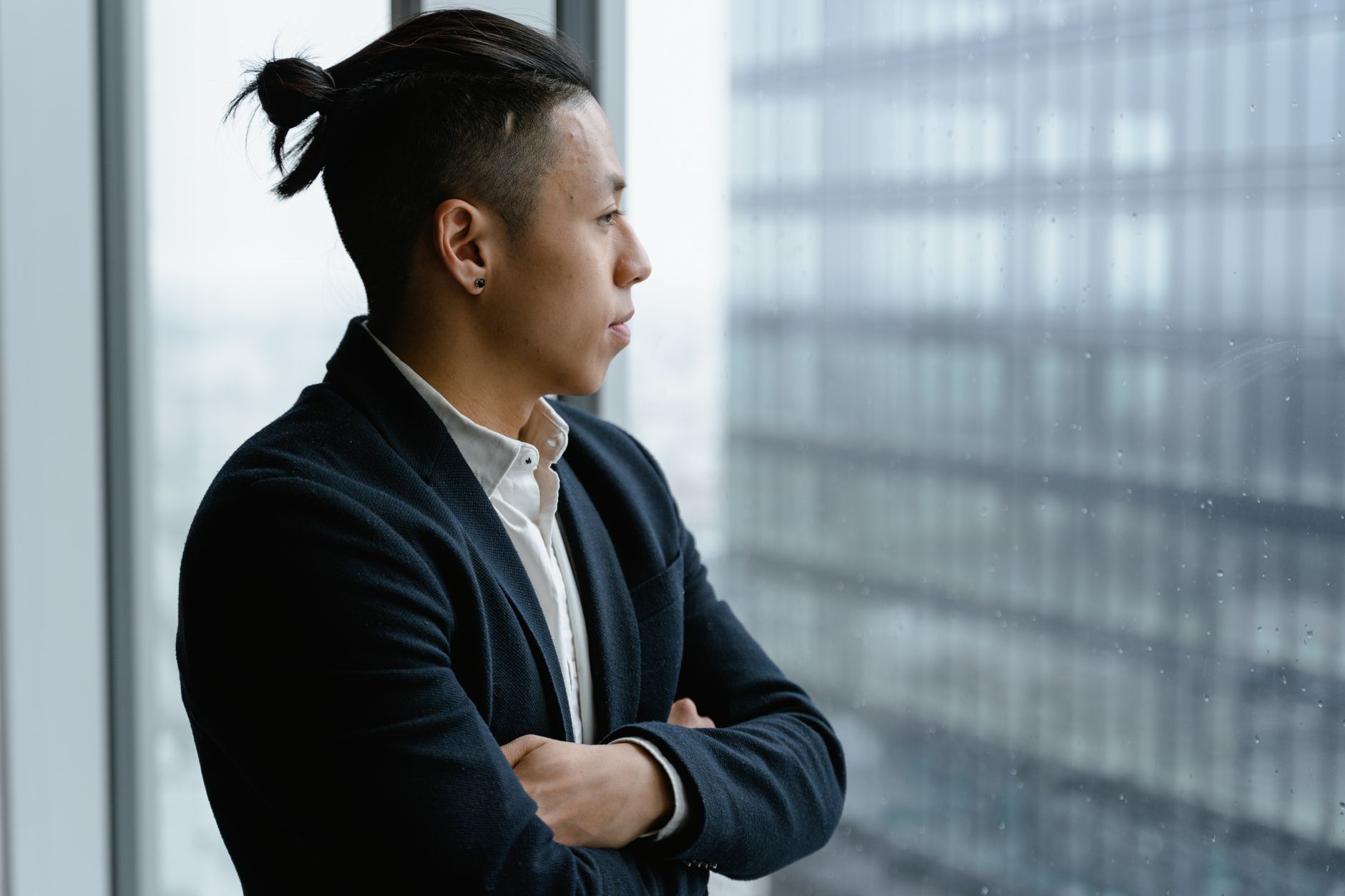 A businessman with arms crossed gazing out of an office window, deep in thought.