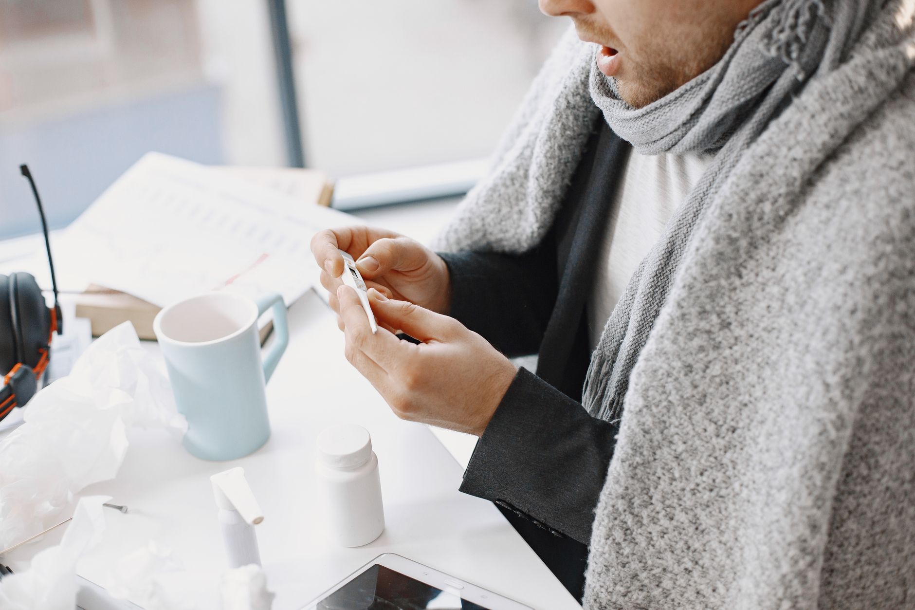 A man wrapped in a blanket, checking a thermometer while convalescing indoors.