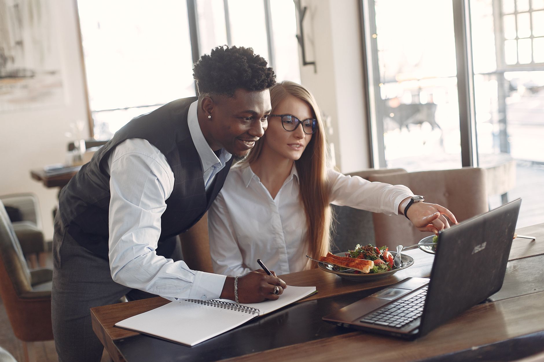 Young diverse entrepreneurs in formal clothes sitting in modern cafe with laptop and planner while working on project together during lunch 