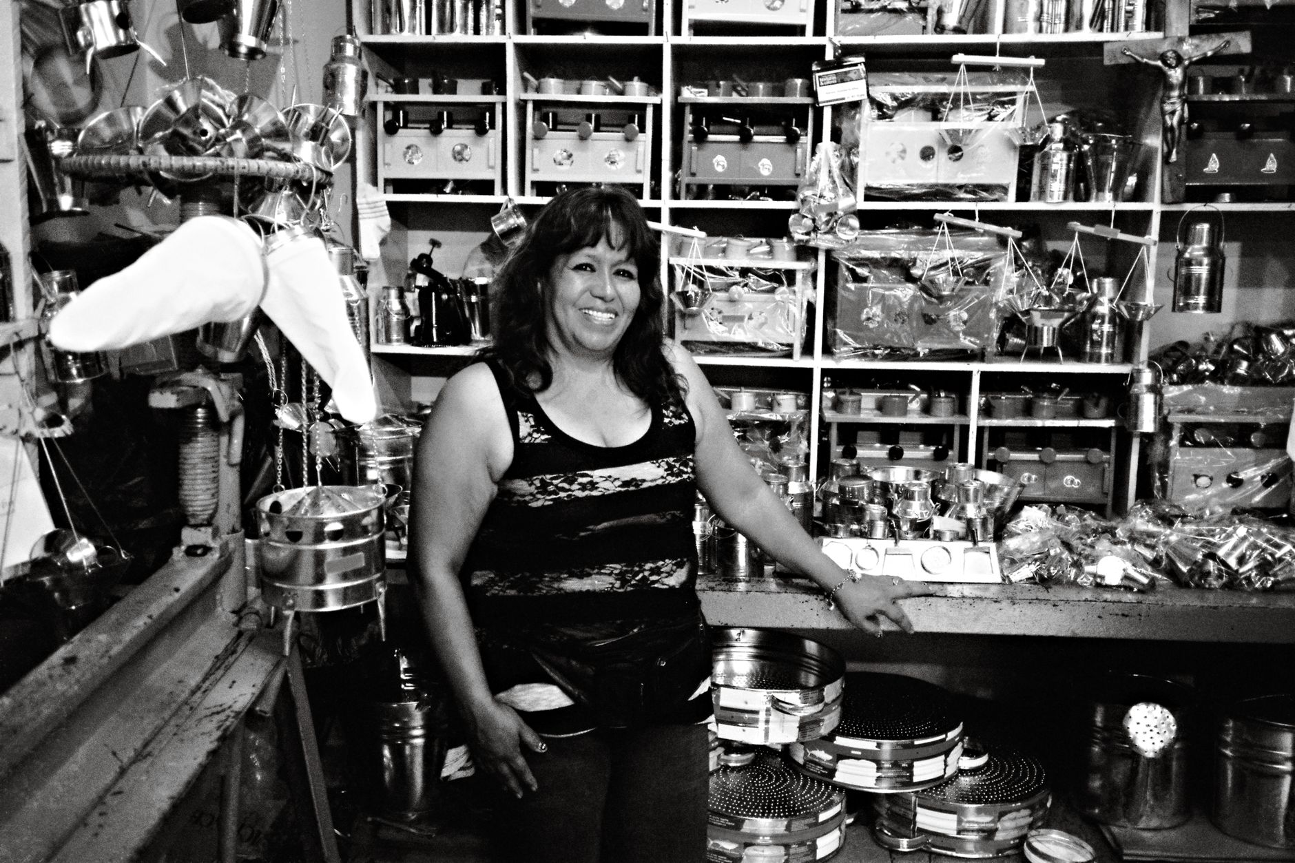 A female shop owner smiling amidst metal goods in a Quito market.