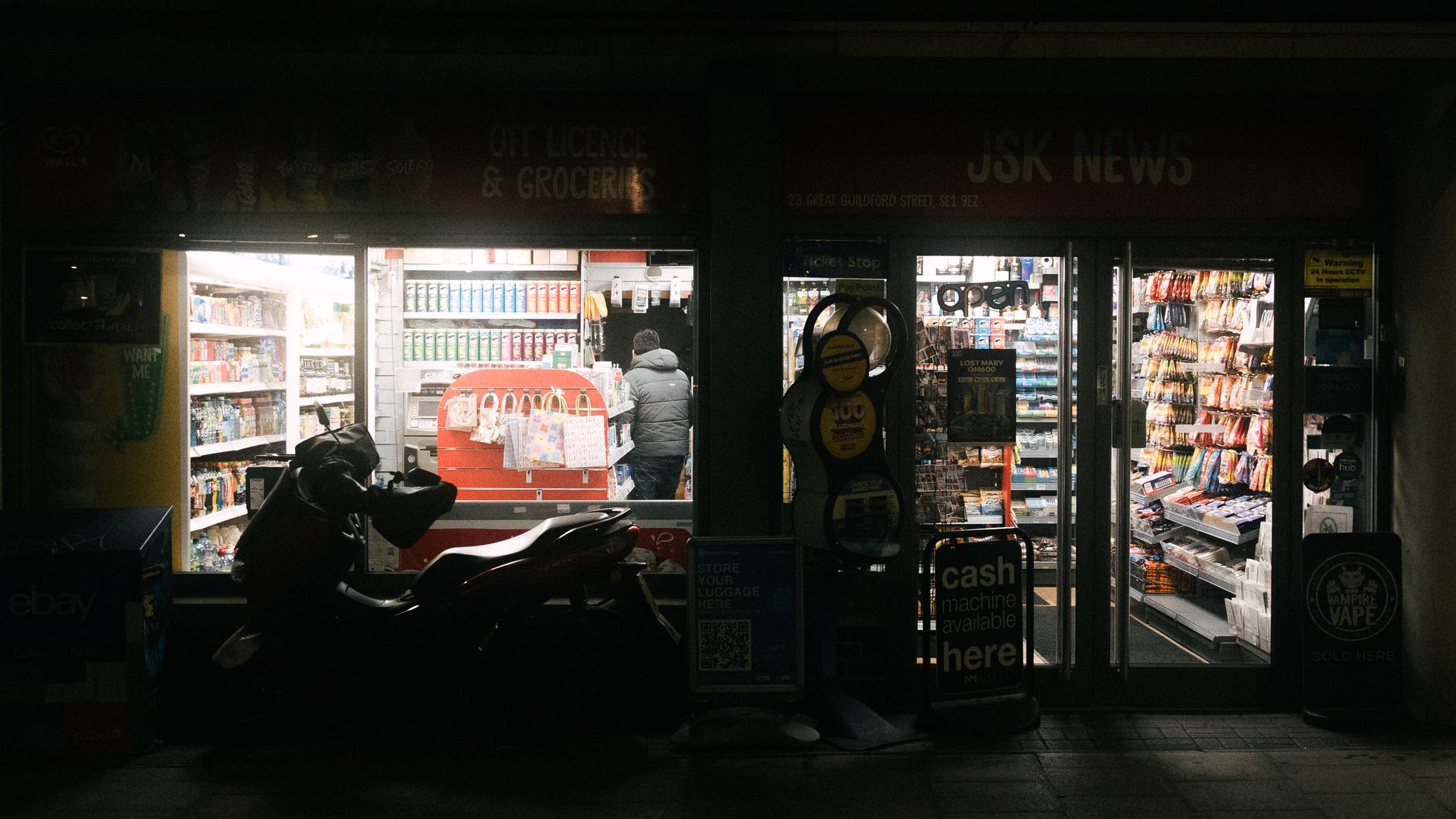 A dimly lit convenience store in London at night with a motorbike parked outside.