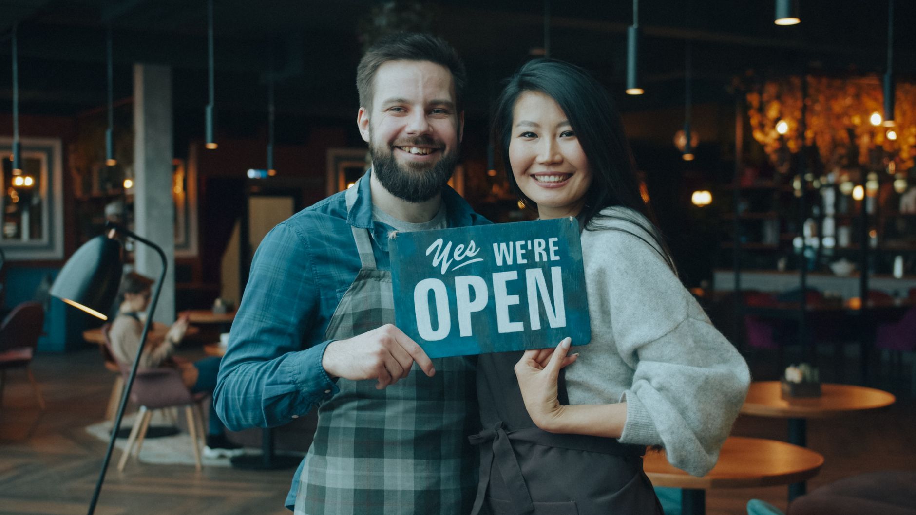 Smiling cafe owners holding an open sign inside their cozy cafe, welcoming customers warmly.