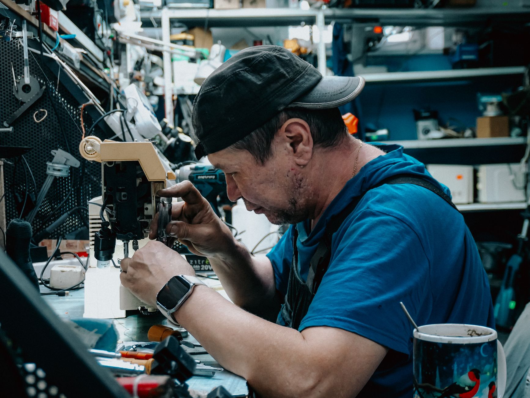 Focused man wearing cap repairs sewing machine in cluttered workshop.